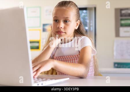 Female child sitting in school working on laptop, resting chin, wearing pink gingham dress Stock Photo