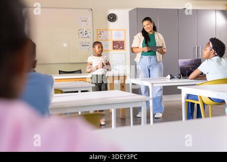 African American woman teacher holding tablet guiding boy speaking in class with laptop, copy space Stock Photo