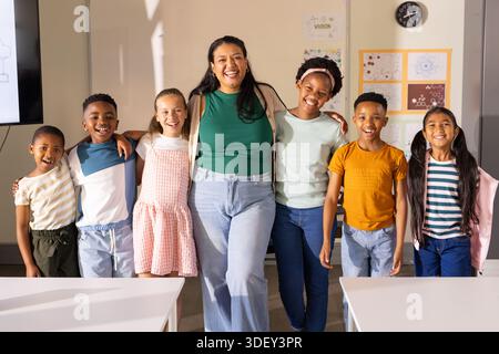 Female teacher hugging children, posing in classroom near white tables and wall display Stock Photo