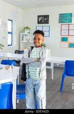 African American boy standing with black backpack slung on shoulder in classroom, headphones nearby Stock Photo