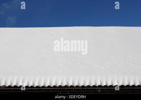 Snow blankets the roof of a building with a clean, blue sky above. The roof shows a smooth white layer. Sunlight shines brightly on the snowy surface. Stock Photo