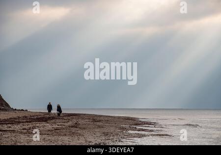 couple walking along a sandy beach in the winter with sunrays and beams of light breaking through the clouds and shining onto the sea. Stock Photo