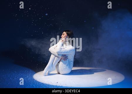 Woman in white sweater dress and knee socks sits against flying snow, cold winter concept, Stock Photo