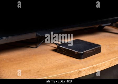 A modern black digital media streaming device connected with cables, sitting on a wood table in front of a flat screen TV. Home entertainment. Stock Photo