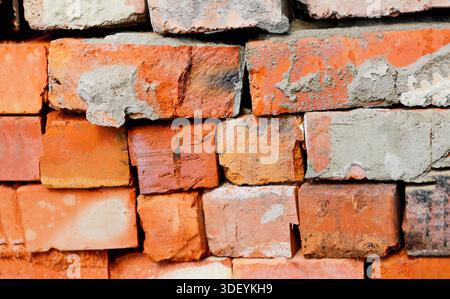 A close-up view of a rough, weathered brick wall. The red and orange clay bricks are stacked unevenly, with visible patches of grey mortar and cement Stock Photo
