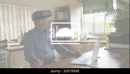 Interacting senior man wearing VR headset, casual shirt using laptop with cloud overlay at kitchen Stock Photo