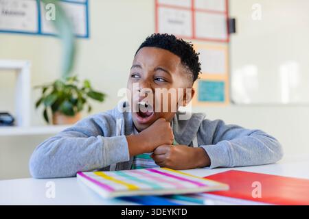 African American preteen in gray hoodie yawning, leaning on desk in classroom with bright notebooks Stock Photo