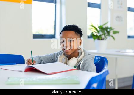 African American male in gray hoodie writing in notebook at school desk with pencil and headphones Stock Photo