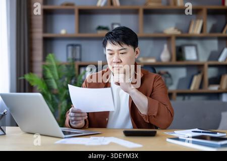Man checking bills and preparing taxes at his home office, analyzing financial documents and using a laptop and calculator for budgeting and debt mana Stock Photo