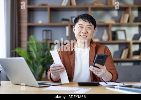 Asian man smiling and holding a receipt and smartphone while sitting at a desk with a laptop and calculator, organizing personal finances and taxes at Stock Photo