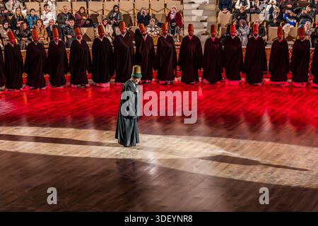 After the end of the Şeb-i Arus ceremony at the Mevlana Kültür Merkezi in Karatay, Konya, Turkey, a venue that holds up to 3,000 people, the dervishes are once again wrapped in their black cloaks, and the Şeyh (sheikh/master) strides towards the exit. The Şeyh, identifiable by his green robe and turban, walks alone across the lit floor in front of the kneeling row of dervishes. Şeb-i Arus is the annual festival marking the death anniversary (17 December) of the mystic poet Rumi (Jalāl ad-Dīn ar-Rūmī, 1207–1273), founder of the Mevlevi order, celebrated as a spiritual union with God. Stock Photo