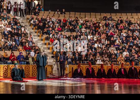 The end of the Şeb-i Arus ceremony at the Mevlana Kültür Merkezi in Karatay, Konya, Turkey, a venue that holds up to 3,000 people, is heralded by a recitation from the Koran. After their whirling dance, the dervishes are once again cloaked in their black robes. A dervish kneels with his hands folded before his face on the red carpet, while a master of ceremony stands beside him and the applauding audience is visible in the background. Şeb-i Arus is the annual festival marking the death anniversary (17 December) of the mystic poet Rumi (Jalāl ad-Dīn ar-Rūmī, 1207–1273), founder of the Mevlevi o Stock Photo