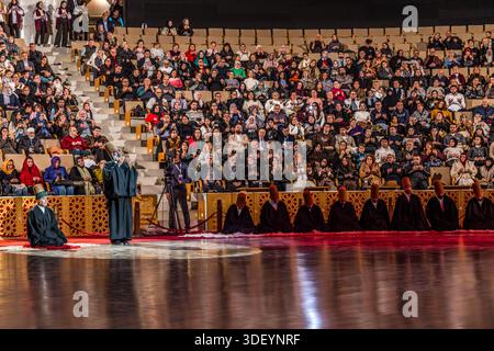 The end of the Şeb-i Arus ceremony at the Mevlana Kültür Merkezi in Karatay, Konya, Turkey, a venue that holds up to 3,000 people, is heralded by a recitation from the Koran. After their whirling dance, the dervishes are once again cloaked in their black robes. A dervish kneels on the red carpet, with two masters of ceremony standing beside him, hands folded, while behind them a long row of dervishes, already dressed in black again, kneels as the audience applauds. Şeb-i Arus is the annual festival marking the death anniversary (17 December) of the mystic poet Rumi (Jalāl ad-Dīn ar-Rūmī, 1207– Stock Photo