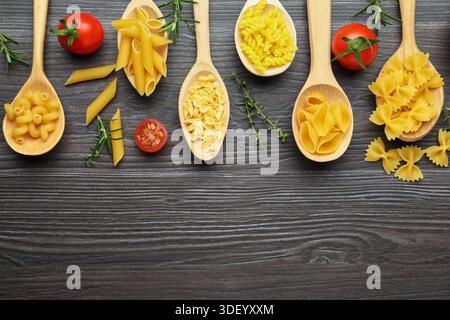 Different types of raw pasta in spoons, tomatoes, rosemary and thyme on wooden table, flat lay. Space for text Stock Photo