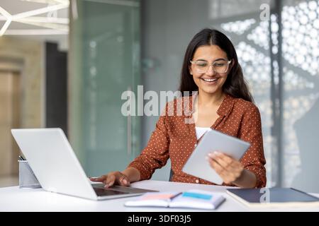 Young happy indian woman working efficiently in a modern office, simultaneously using a laptop and a digital tablet, reflecting productivity and innov Stock Photo