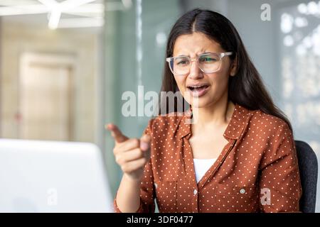 Angry young woman wearing glasses arguing and pointing her finger at a laptop screen during a virtual meeting, expressing frustration and disagreement Stock Photo