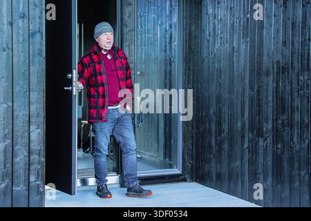 Elderly man in plaid shirt and beanie stands at doorway, highlighting modern wooden architecture and inviting ambiance of home, emphasizing warmth and Stock Photo