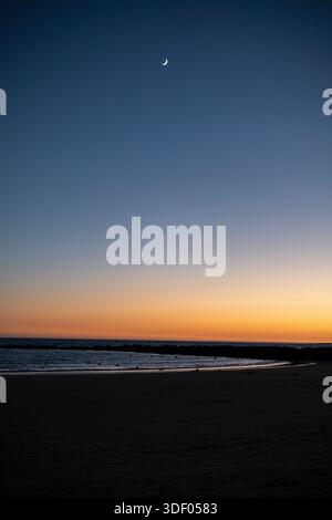 Crescent moon illuminating the sky above a calm beach with an orange and blue sunset gradient Stock Photo