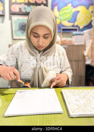 Young woman in hijab working on Islamic Batik geometric art with a traditional tool at a creative studio desk workshop Stock Photo