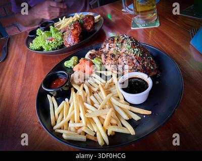 Grilled chicken served with French fries, steamed vegetables, and dipping sauces on a black plate at a restaurant table Stock Photo