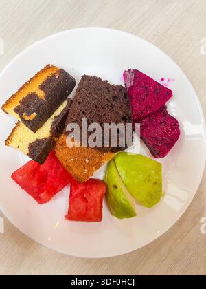 Assorted slices of cake and fresh tropical fruit including watermelon, dragon fruit, and guava on a white plate malaysia Stock Photo