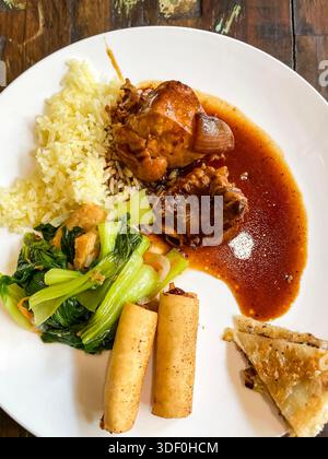Assorted Asian-style meal with braised chicken in sauce, rice, spring rolls, sautéed greens, and pastry on a white plate Stock Photo