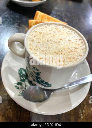 A cup of frothy ipoh white coffee  ot Teh C served in a traditional patterned ceramic mug  on a wooden table Stock Photo