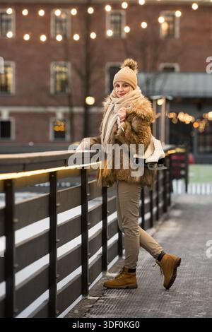 Smiling young woman standing near ice rink holding white skates, looking at camera. Winter leisure Stock Photo