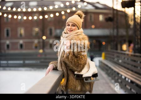 Smiling young woman standing near ice rink holding white skates, looking at camera. Winter leisure Stock Photo
