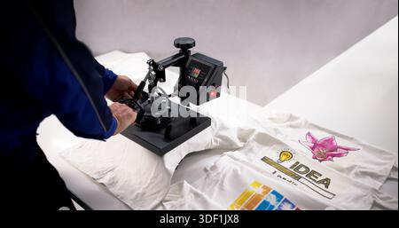 Young Hispanic Man Using Heat Press Machine To Print Custom Shirt Logo. Stock Photo