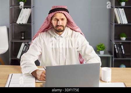 Focused businessman in traditional attire working on laptop at modern office desk Stock Photo