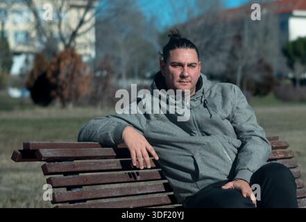 Man with man bun sitting calmly on a public park bench outdoors Stock Photo