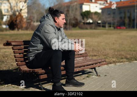 Young man sitting on a park bench outdoors, holding a cigarette and thinking Stock Photo