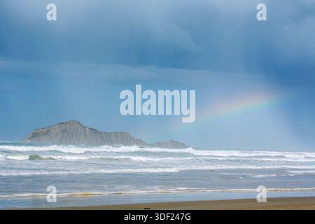 A scenic view of a beach with waves crashing against the shore, featuring a distant rocky island under a cloudy sky. A rainbow arcs over the ocean, ad Stock Photo