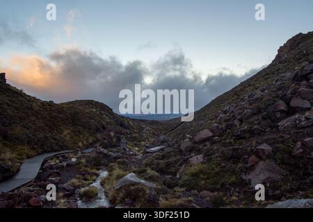 A scenic view of a valley between two hills, with a wooden pathway winding through rocky terrain. The sky is partly cloudy, with soft hues of orange a Stock Photo