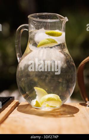 A clear glass pitcher filled with cold water, ice cubes, and lime slices stands on a wooden surface in bright sunlight. Stock Photo