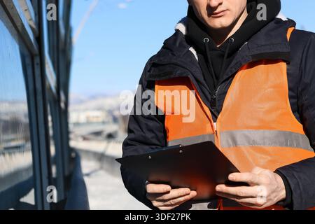 Person in high-visibility orange vest and dark jacket examines documents on clipboard against snowy mountain vista Stock Photo
