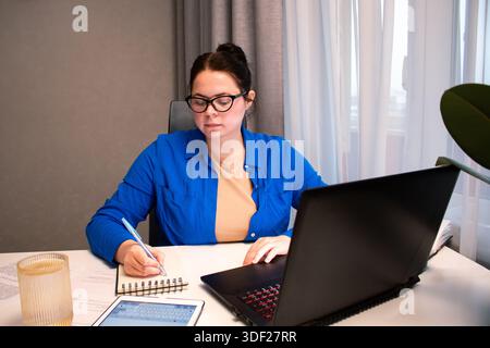 Student attending virtual university class via video conference. Young woman actively participates in online lecture, taking notes on laptop. Distance Stock Photo