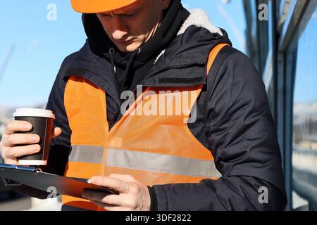Male in hard hat holding hot drink, looking at smartphone over clipboard on snowy overpass overlooking city and snowy mountains. Stock Photo
