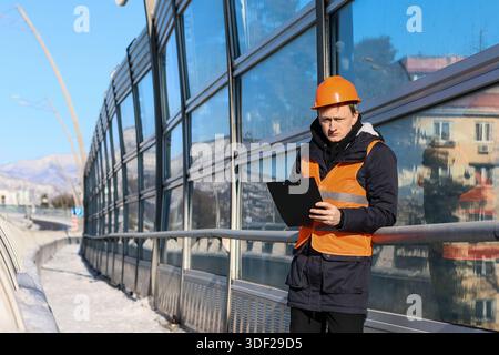 Engineer in orange safety attire leans on railing, reading notes near transparent barrier reflecting cityscape in winter conditions. Stock Photo