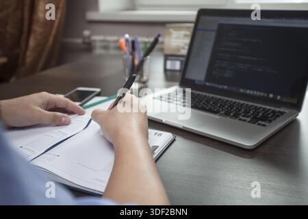 Man's hands make entries in the notebook, an open laptop in the face of Stock Photo