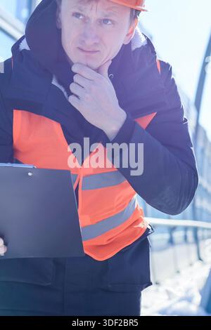 Worker in orange safety vest ponders while holding clipboard on sunny construction site Stock Photo
