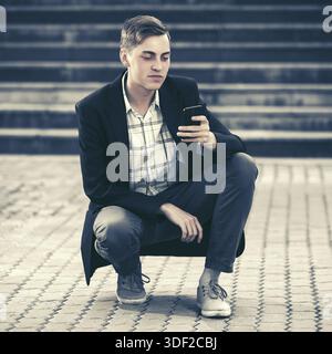Young handsome business man using smart phone sitting in city street. Stylish trendy male model in black jacket Stock Photo