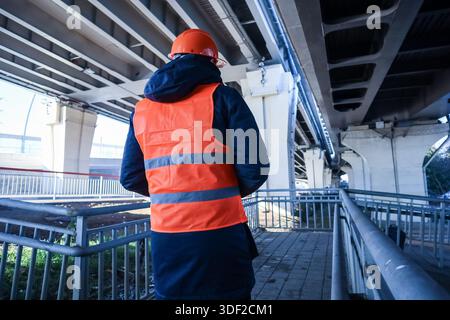 Silhouette of engineer in high-visibility attire inspecting pillars and framework beneath elevated highway. Stock Photo
