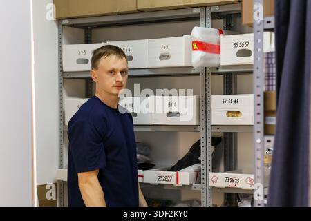 Man in blue shirt poses neutrally by metal racks filled with white boxes and various items. Stock Photo