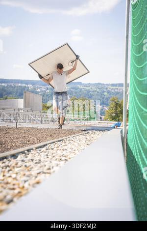 Man carrying a solar panel across a building site with a view and green fence, new park and ride parking garage Calw Heumaden on the new Hermann Hesse Stock Photo