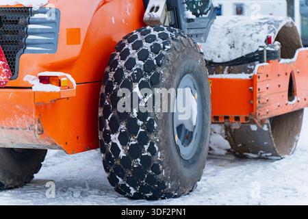 Close up of orange road roller wheel with textured rubber tire and snow buildup showing heavy construction machinery traction and winter roadwork cond Stock Photo