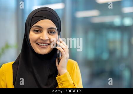 Smiling young muslim businesswoman in a black hijab and yellow blouse, confidently talking on a cellphone in a modern office, conveying professionalis Stock Photo