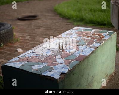 Mosaic park bench with a small metal item on the surface, suggesting a found object in a public outdoor space. Stock Photo