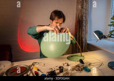 Man crafter renovating vintage lamp at home workshop, sitting at table assembling item parts, screwing in lampshade to original base holder using screws, checking fixation, upcycling and refurbishing. || Model released Stock Photo